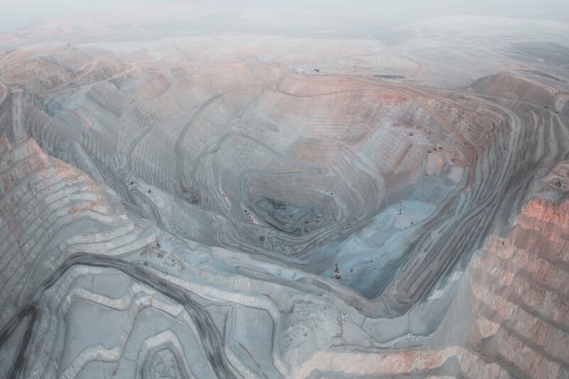 Aerial view of a large open-pit mine with terraced sides and winding roads. The landscape is arid with a mix of gray and reddish-brown hues, indicating different mineral layers. A few vehicles are visible inside the pit.