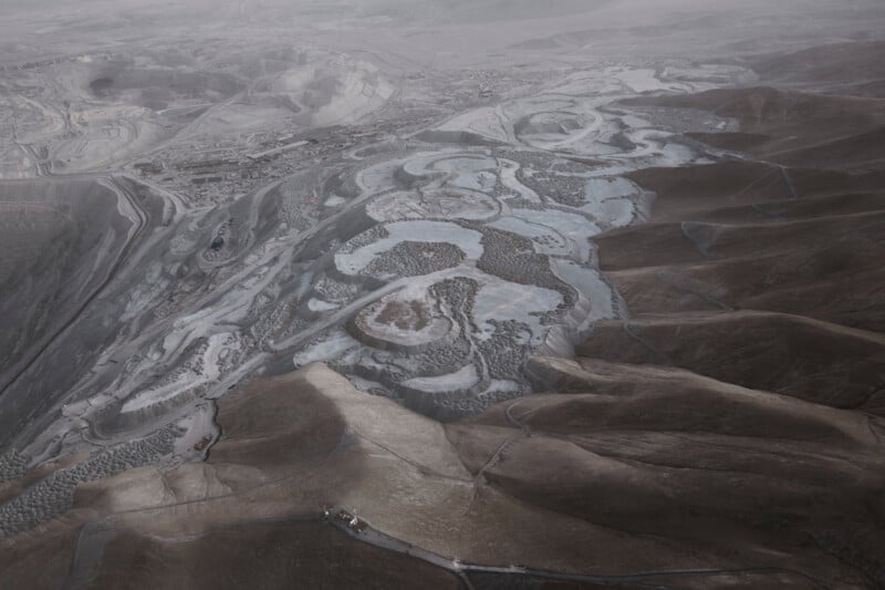 Aerial view of a rugged landscape featuring winding roads and pathways cutting through hills. The scene appears desolate and muted, with shades of brown and gray dominating the terrain. Subtle patterns and textures create an abstract visual effect.