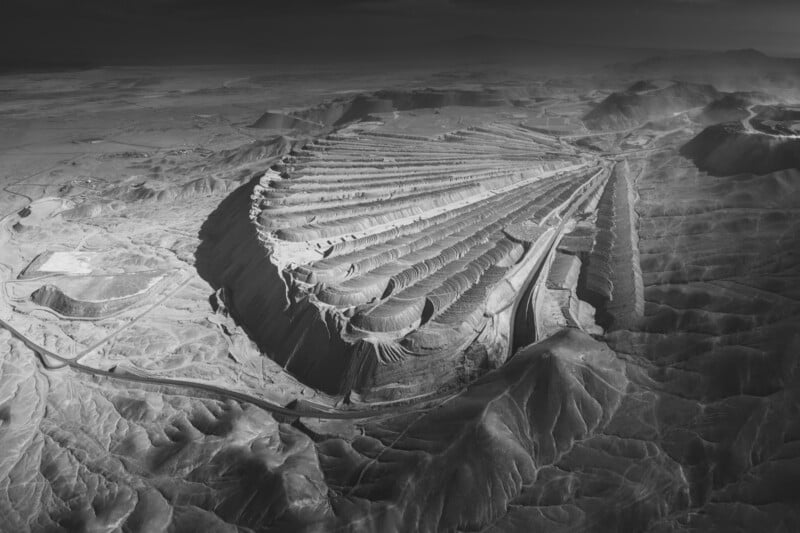 A vast aerial view of an open-pit mine with terraced layers, creating a dramatic, geometric landscape. Surrounding the mine are barren, undulating hills and a few roads, all rendered in grayscale, emphasizing the texture and scale.