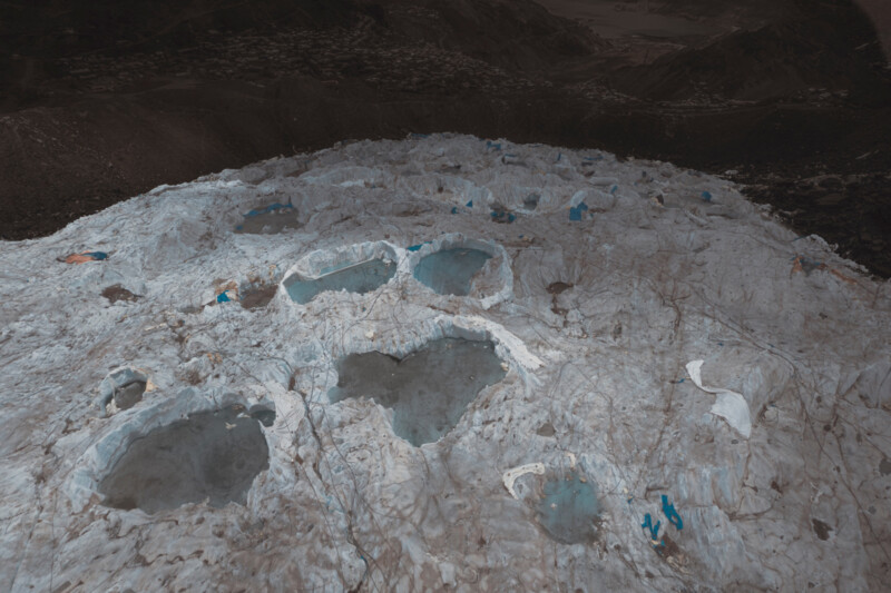 Aerial view of a snowy glacier with multiple large, blue melt ponds scattered across its surface. Small figures in bright clothing are visible around the ponds, emphasizing the vastness of the icy landscape. Dark mountains loom in the background.