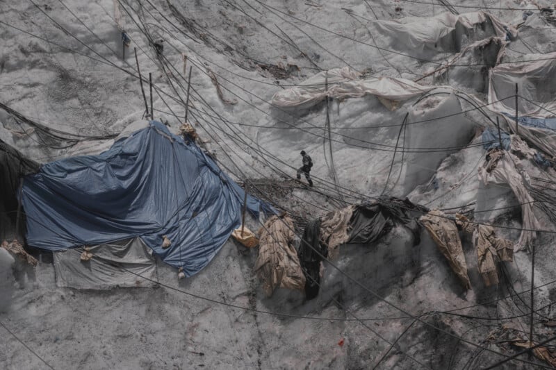 A person stands amidst a glacier, surrounded by a network of ropes and tarps in blue and beige. The icy surface is uneven, with various peaks and patches. The scene conveys a sense of survival or exploration in a harsh, frigid environment.