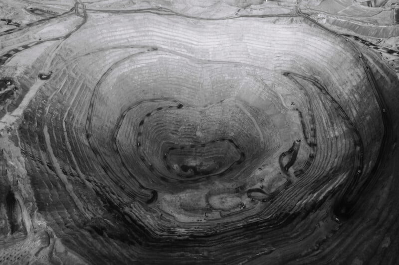 Aerial view of a large, circular open-pit mine with terraced layers. The spiraling design creates a pattern resembling a giant crater. Various pathways and roads are visible along the layers, and the landscape appears barren. Image in black and white.