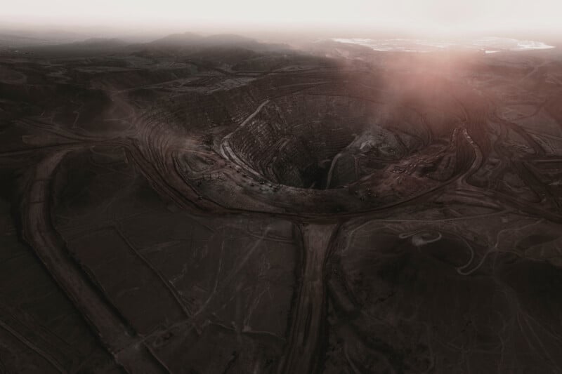 Aerial view of a large, circular open-pit mine with terraced edges, surrounded by barren land and faint road networks. The scene is bathed in a reddish hue, with mist and a sunlit haze in the distant sky.