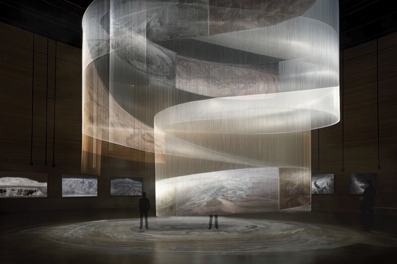 A dimly lit exhibition space features a large, translucent spiral installation hanging from the ceiling. People stand below, observing the layered structure, with framed images lining the wooden walls in the background.