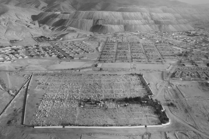 Aerial view of a vast desert landscape; foreground shows a large, rectangular cemetery with numerous grave markers. The area is enclosed by a wall. In the background, residential buildings and barren mountains are visible. Image is in black and white.