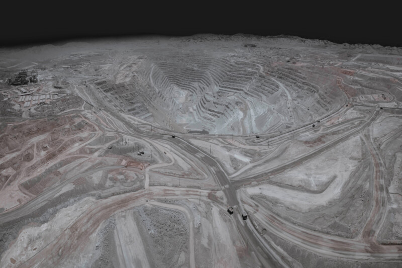 Aerial view of a large, terraced open-pit mine under a cloudy sky. Various dirt roads connect the levels, and several trucks are visible on the paths. The landscape is barren with varying shades of brown and gray.