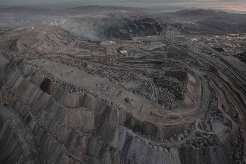 Aerial view of a vast, barren landscape featuring large terraced layers of an open-pit mine. The terrain is brown and dusty, with winding roads and scattered equipment. Mountains and a cloudy sky can be seen in the distance.