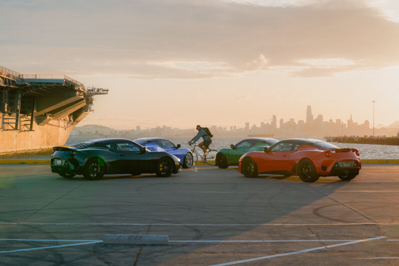 Four sports cars—gray, blue, green, and red—are parked in a semicircle on a waterfront, with a cyclist riding nearby. In the background, a city skyline and a partly cloudy sky are visible during sunset.
