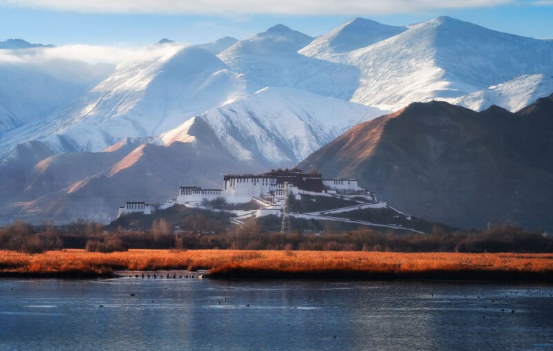 A majestic, white palace sits amidst a stunning landscape of snow-capped mountains and vibrant autumn-colored foliage. A peaceful lake in the foreground reflects the scenery, adding to the serene and picturesque view.