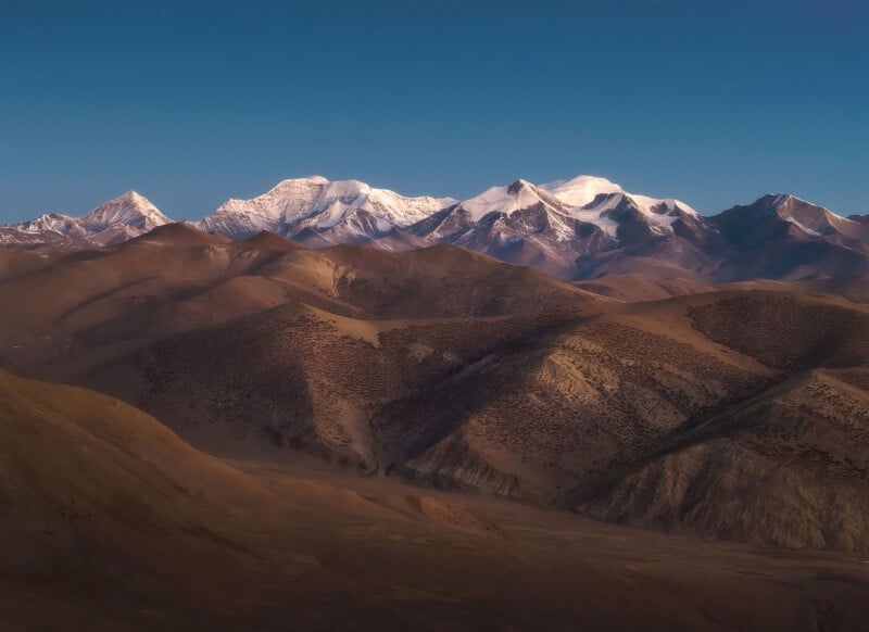 Mountain range with snow-capped peaks under a clear blue sky. The foreground features rugged brown hills with sparse vegetation. The sunlight casts soft shadows, highlighting the texture of the landscape.