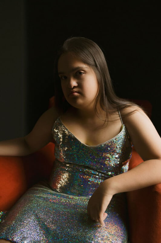 A person with long hair sits in a red chair, wearing a shimmering sequin dress. They have a calm and confident expression. The background is dark, highlighting the individual in the seat.