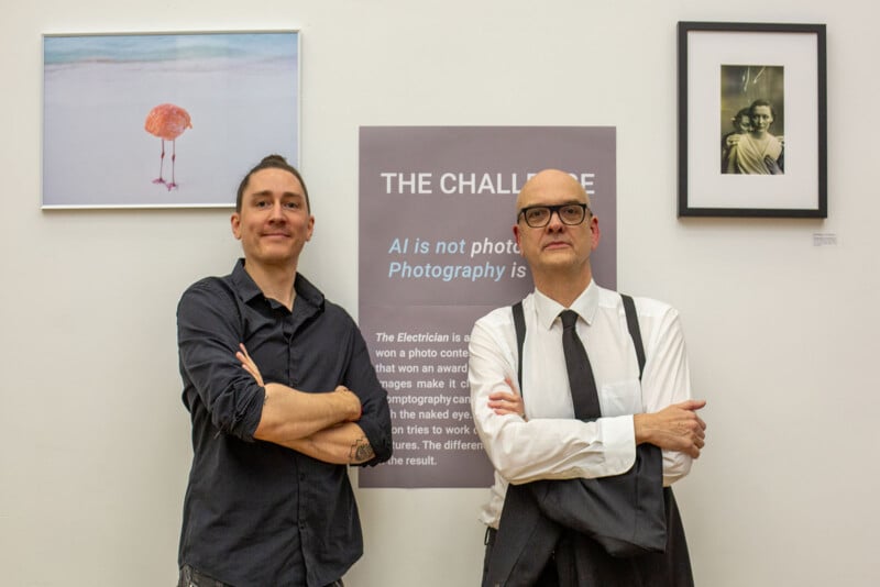 Two men stand with arms crossed in front of a wall displaying framed photographs and a poster titled "THE CHALLENGE: AI is not photography." One man wears a dark shirt, the other a white shirt with a tie and suspenders.