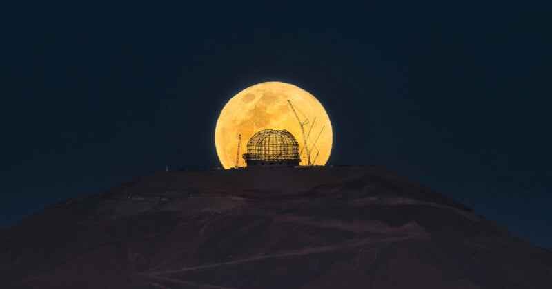 A large, full moon rises in the night sky over a silhouetted observatory dome on a hilltop. Construction cranes are visible around the dome, creating a striking scene against the dark background.