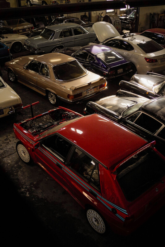 A cluttered garage filled with various cars, including a prominent red car with a spoiler and racing stripes. Some cars have their hoods open, and there's a mix of classic and modern vehicles, all packed closely together.