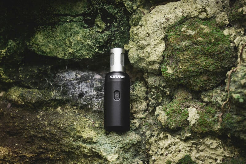 A black Shure microphone is leaning against a textured, moss-covered stone wall. The contrast highlights the sleek design of the microphone against the natural, rugged surface.