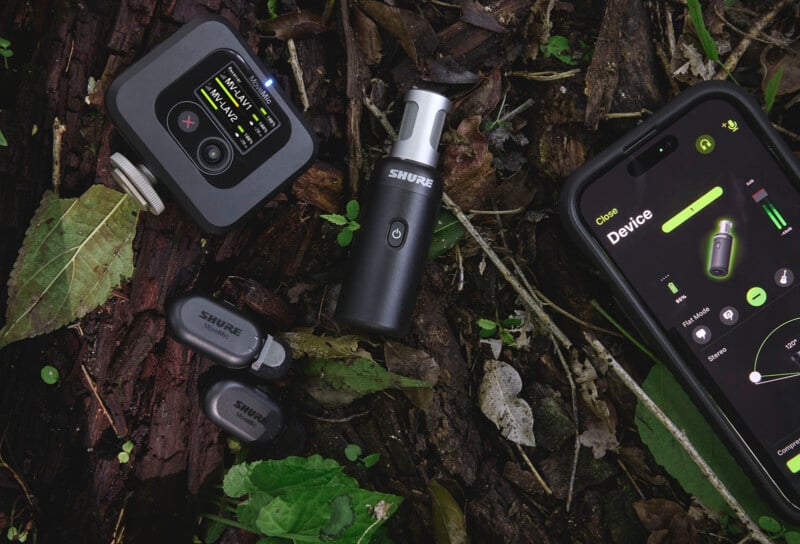 Audio equipment on forest ground: Shure transmitter, receiver, earbuds, and a smartphone showing device interface. Placed on earth with leaves and bark.
