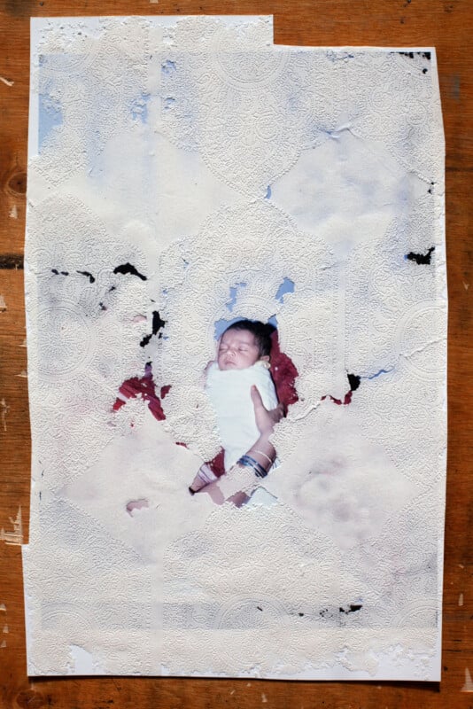 A photograph of an infant wrapped in red cloth is pasted onto a textured white paper background with floral patterns. The paper appears worn and partially torn, revealing areas of the image beneath.