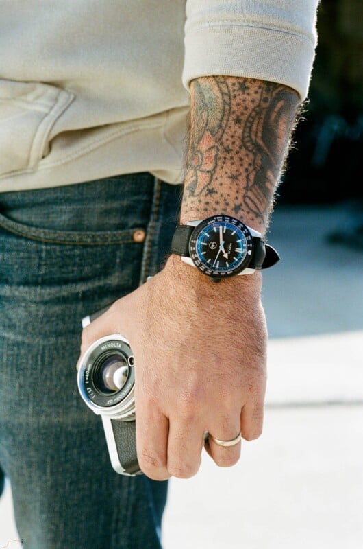 A person with a tattooed arm wears a watch with a blue and black face, holding a vintage Minolta camera. The person is dressed in jeans and a light-colored sweater.