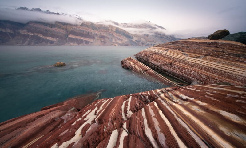 A serene landscape featuring red and white striped rock formations beside a calm, turquoise lake. In the background, foggy mountains rise under a cloudy sky, adding depth to the tranquil scene.