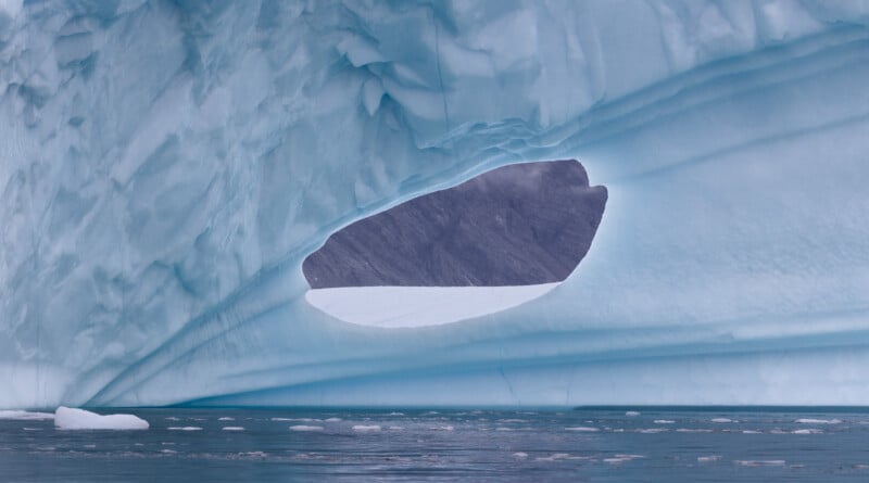 A close-up of a blue iceberg with a smooth, oval-shaped opening revealing a view of distant gray mountains and cloudy sky. The iceberg's texture is visible, with ripples and layers, above the calm, icy water.