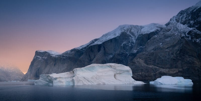 A serene landscape features large icebergs floating in calm water, surrounded by towering, snow-capped mountains under a pastel-colored sky at dusk.