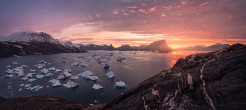 A scenic view of an Arctic landscape at sunrise. Icebergs float in a calm, dark blue sea surrounded by jagged mountains. The sky is painted with warm hues of orange and pink, creating a glowing horizon. Rocky foreground adds depth to the scene.