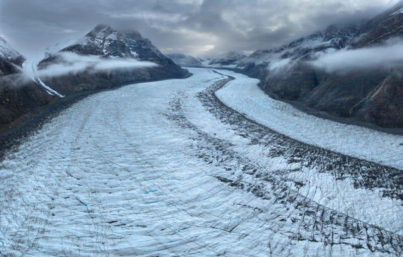 Aerial view of a vast glacier winding through a mountainous landscape under a cloudy sky. Snow-capped peaks flank the icy expanse, with patches of fog hovering around the mountain tops. The glacier has visible crevices and jagged textures.