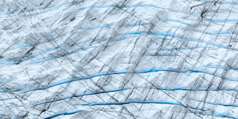 A close-up view of a glacier surface, displaying layers of bluish ice with thin, dark lines and crevices. The texture appears rough, with varying shades of white and blue, highlighting the natural patterns of the ice.