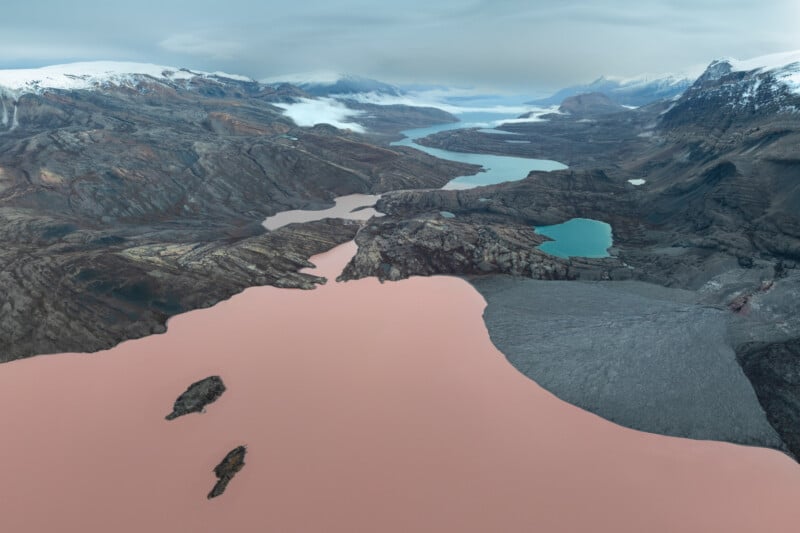 Aerial view of glacial terrain with a striking contrast between a reddish-brown lake and a turquoise lake. Rugged mountains and snow-capped peaks surround the lakes under a cloudy sky, creating a dramatic landscape.