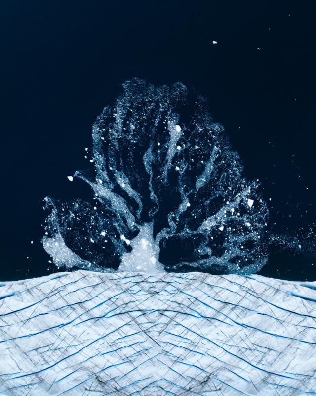 A large splash of water erupts above the surface of a deep blue ocean, creating a tree-like shape. Ice or snow fragments are scattered around, contrasting with the dark water. The lower part shows textured, icy surfaces.