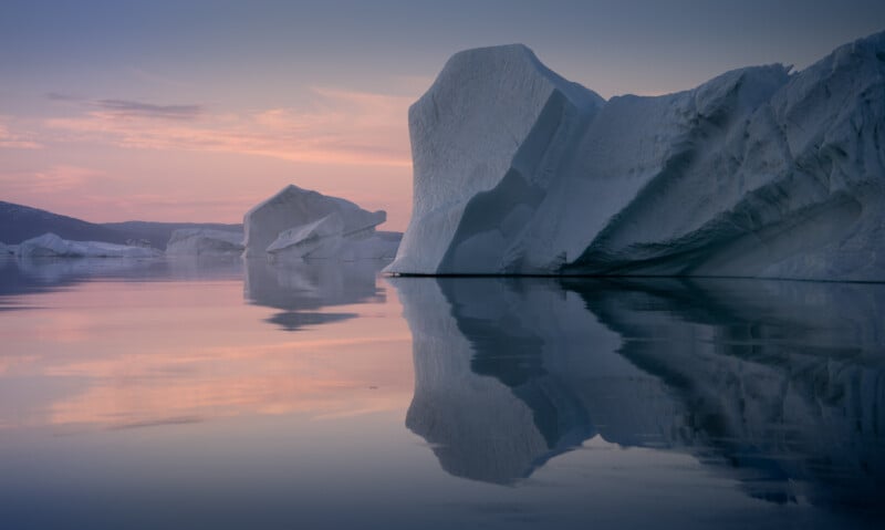 A serene scene of large icebergs floating in calm waters under a pink and blue sunset sky. The icebergs are mirrored in the reflective surface of the water, creating a peaceful and picturesque landscape.