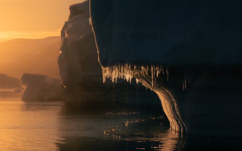 Icebergs in a calm ocean at sunset, with delicate icicles hanging from the foreground iceberg. Warm golden light reflects on the water, creating a serene and majestic atmosphere.