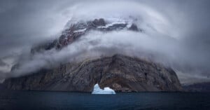 A dramatic mountain partially obscured by thick, misty clouds rises above a dark body of water. A small iceberg floats in the foreground, contrasting against the rocky mountain and cloudy sky.