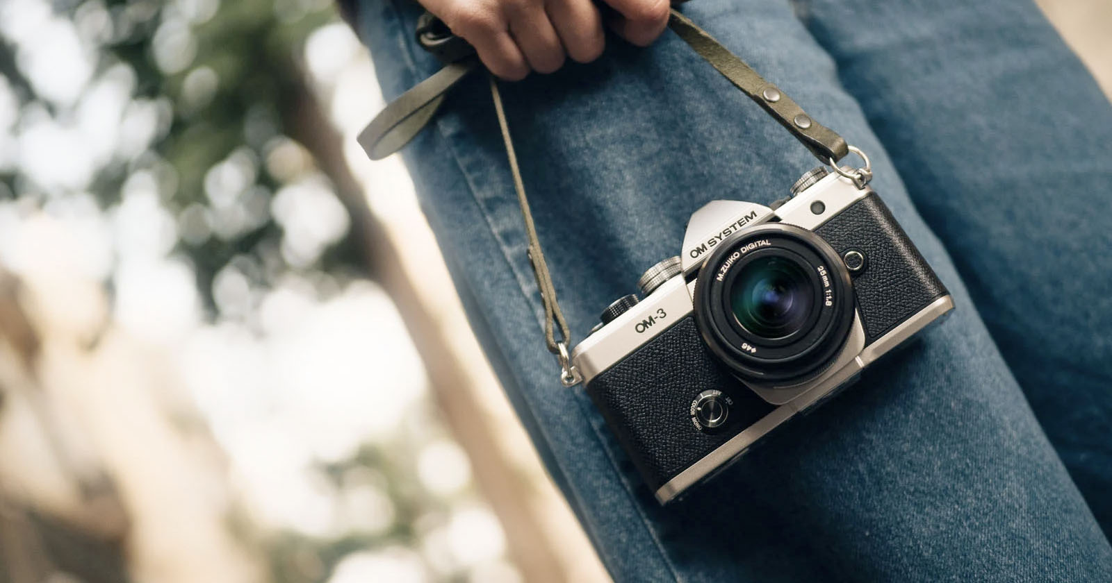 Close-up of a person holding a vintage-style camera by its strap, with green trees blurred in the background. The person is wearing blue jeans, and the focus is on the camera lens.