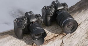 Two Olympus cameras with attached lenses are placed on a weathered wooden log. The cameras are angled slightly towards each other, showcasing their controls and logos against a blurred background.