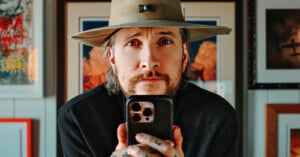 A person wearing a wide-brimmed hat holds a smartphone and looks directly at the camera. The background features framed artwork on the wall.