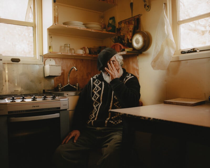 An elderly man sits at a kitchen table, covering his face with one hand. He wears a patterned sweater and a cap. The kitchen features a stove, sink, and hanging pot. Natural light filters through the windows, creating a warm atmosphere.