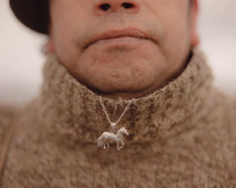 A man wearing a thick knitted turtleneck sweater with a silver horse pendant necklace. Only the lower part of his face is visible, showing a neutral expression. The sweater is textured, and he is also wearing a black beret.