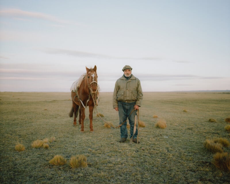 A person in a green jacket and hat stands in an open grassy field beside a brown horse. The sky is clear with few clouds, and sparse vegetation covers the landscape.