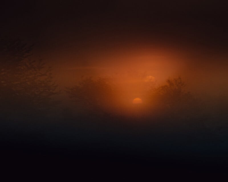 A dark, atmospheric landscape with a vibrant orange glow from the setting sun, partially obscured by silhouette trees and mist. The scene has a mysterious, dreamlike quality, with soft light blending into the dark blue surrounding.