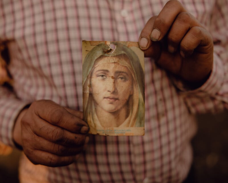 A person in a plaid shirt holds a worn, stained card depicting a religious image of a woman wearing a veil. The hands appear weathered, and the card shows signs of wear and tear.