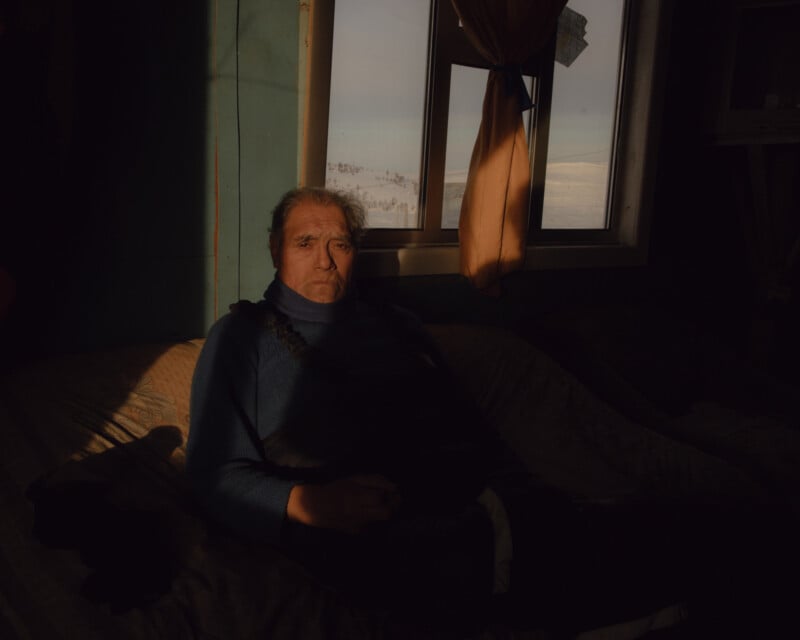 A person sits on a bed in a dimly lit room, with sunlight streaming through a window. They are wearing a blue sweater and dark pants, looking towards the camera. The window curtain is partially open, revealing a snowy landscape outside.