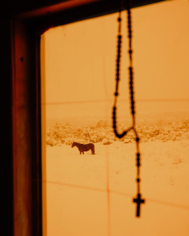 A silhouetted horse stands in a snow-covered field under an orange sky, viewed through a window. In the foreground, a rosary with a cross hangs, creating a tranquil and contemplative scene.