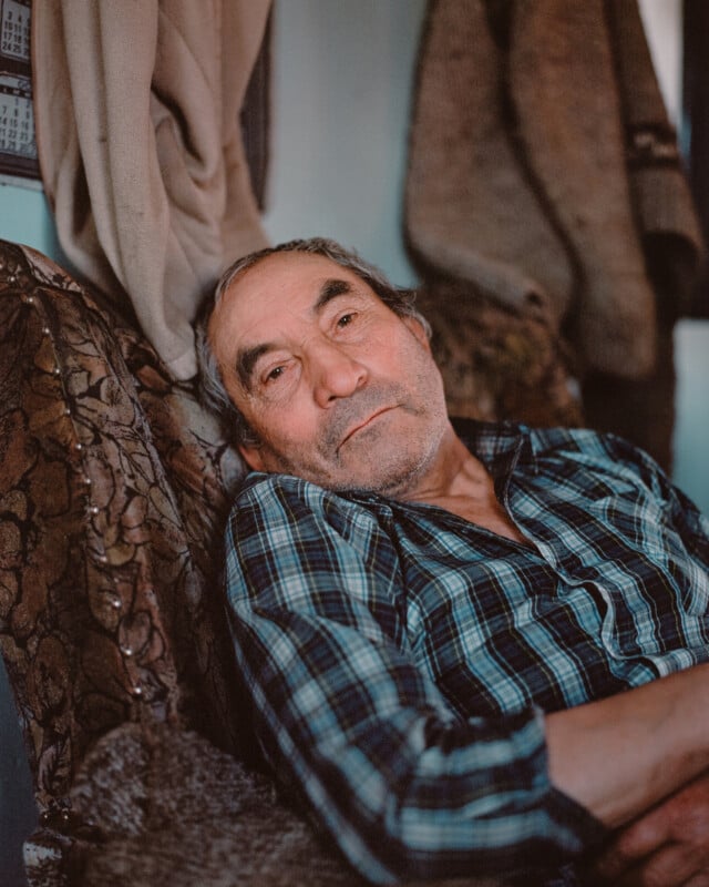 Elderly man with gray hair, wearing a blue plaid shirt, sits on a patterned couch. He appears thoughtful and relaxed. Coats hang nearby, adding warmth to the cozy, softly lit room.