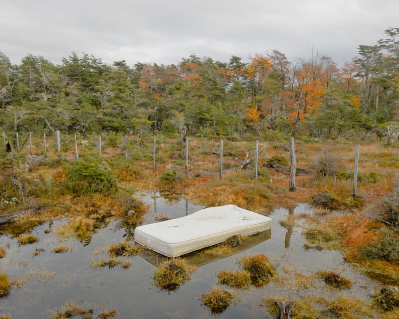 A discarded mattress floats on a wet, marshy area surrounded by sparse vegetation and autumn-colored trees. A simple wire fence is visible in the background under an overcast sky.