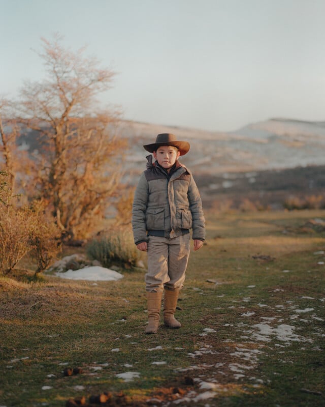 A person wearing a wide-brimmed hat and winter clothing stands on a grassy path in a mountainous landscape. Bare trees and patches of snow are visible, with soft light suggesting early morning or late afternoon.