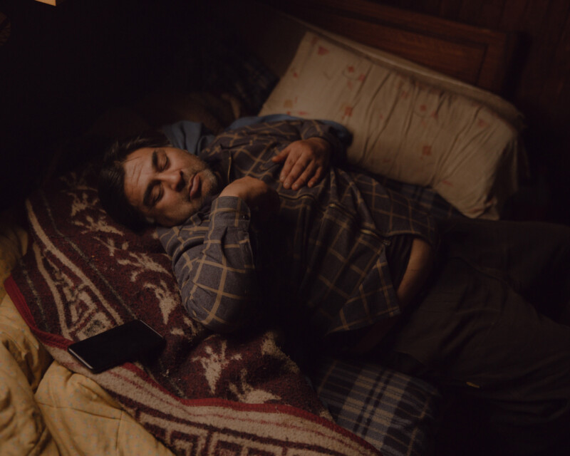 A man is lying on a bed with a patterned blanket, wearing a plaid shirt. His eyes are closed, and a smartphone is next to him. The setting is dimly lit, giving a calm, restful atmosphere.