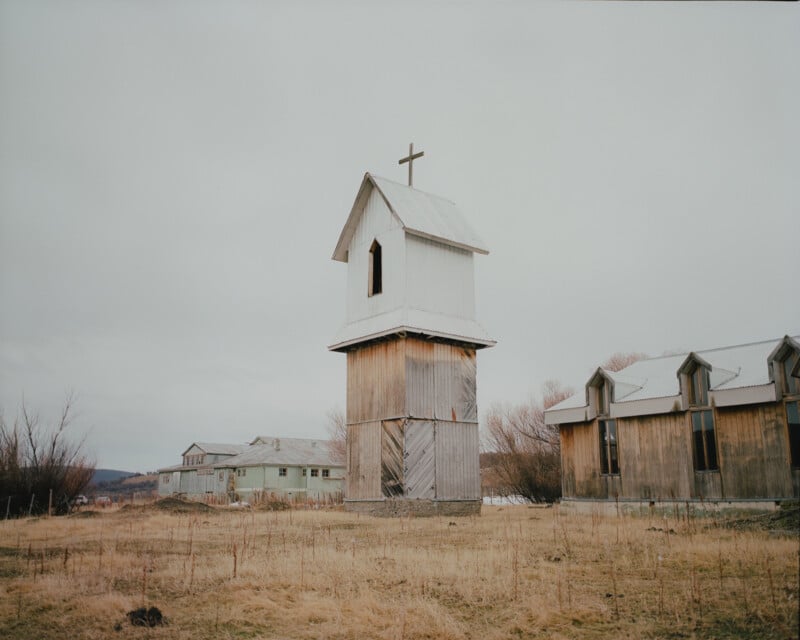 A weathered wooden bell tower with a cross stands in a grassy field under an overcast sky, surrounded by old, rustic buildings. The scene evokes a sense of rural, historic simplicity and tranquility.