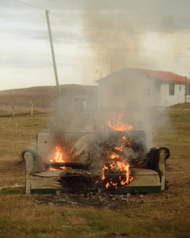 A decrepit couch on a grassy field is engulfed in flames, producing thick smoke. In the background, a small house with a red roof is visible under a cloudy sky.