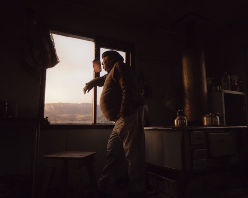 A person in warm clothing stands inside a dimly lit room, gazing out a window with mountains in the distance. There's a wood stove with a kettle and pot, and sunlight streams in, casting a warm glow.